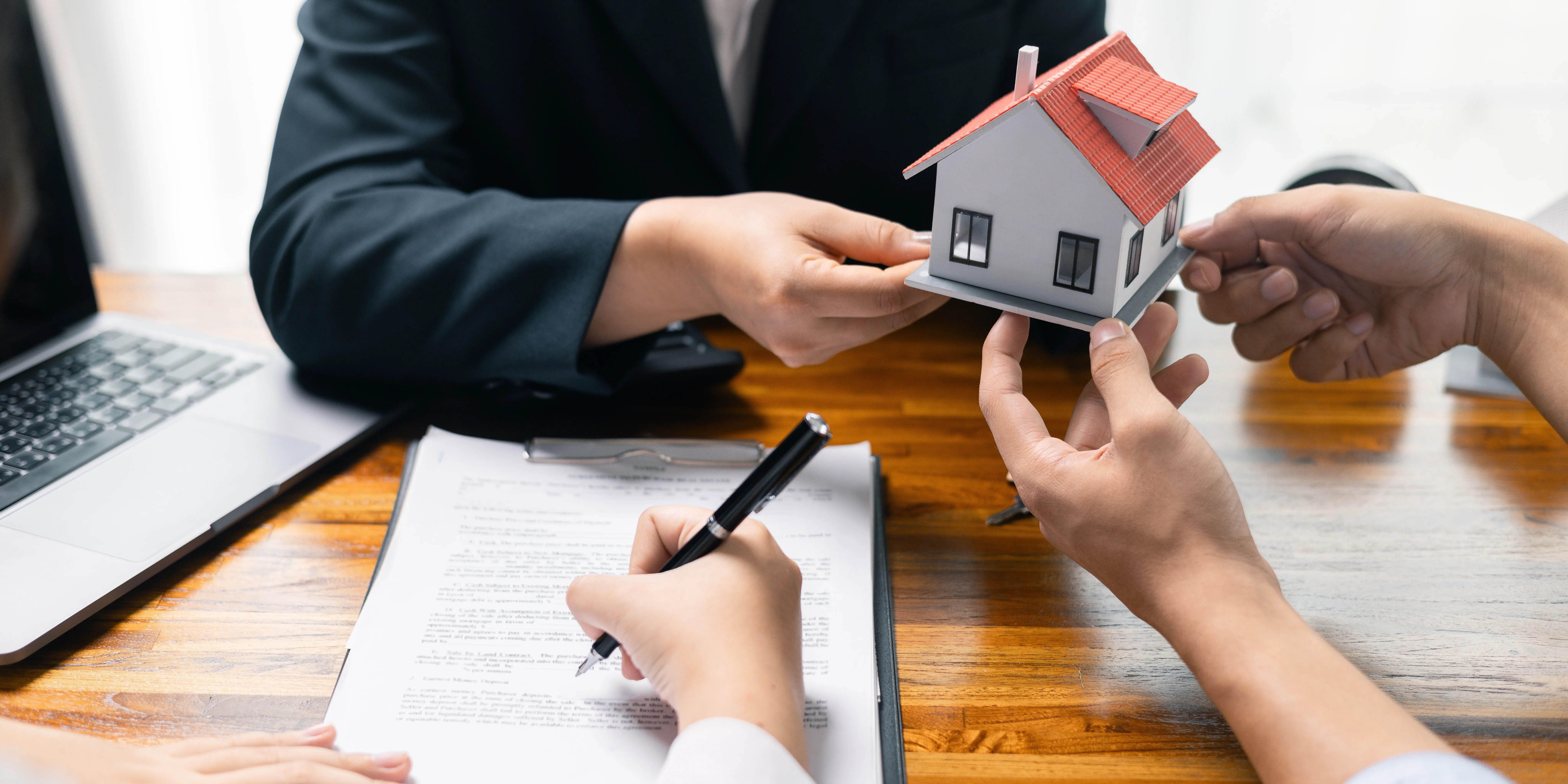 Agente inmobiliario entregando una casa de juguete a una persona mientras que otra persona está firmando el crédito hipotecario