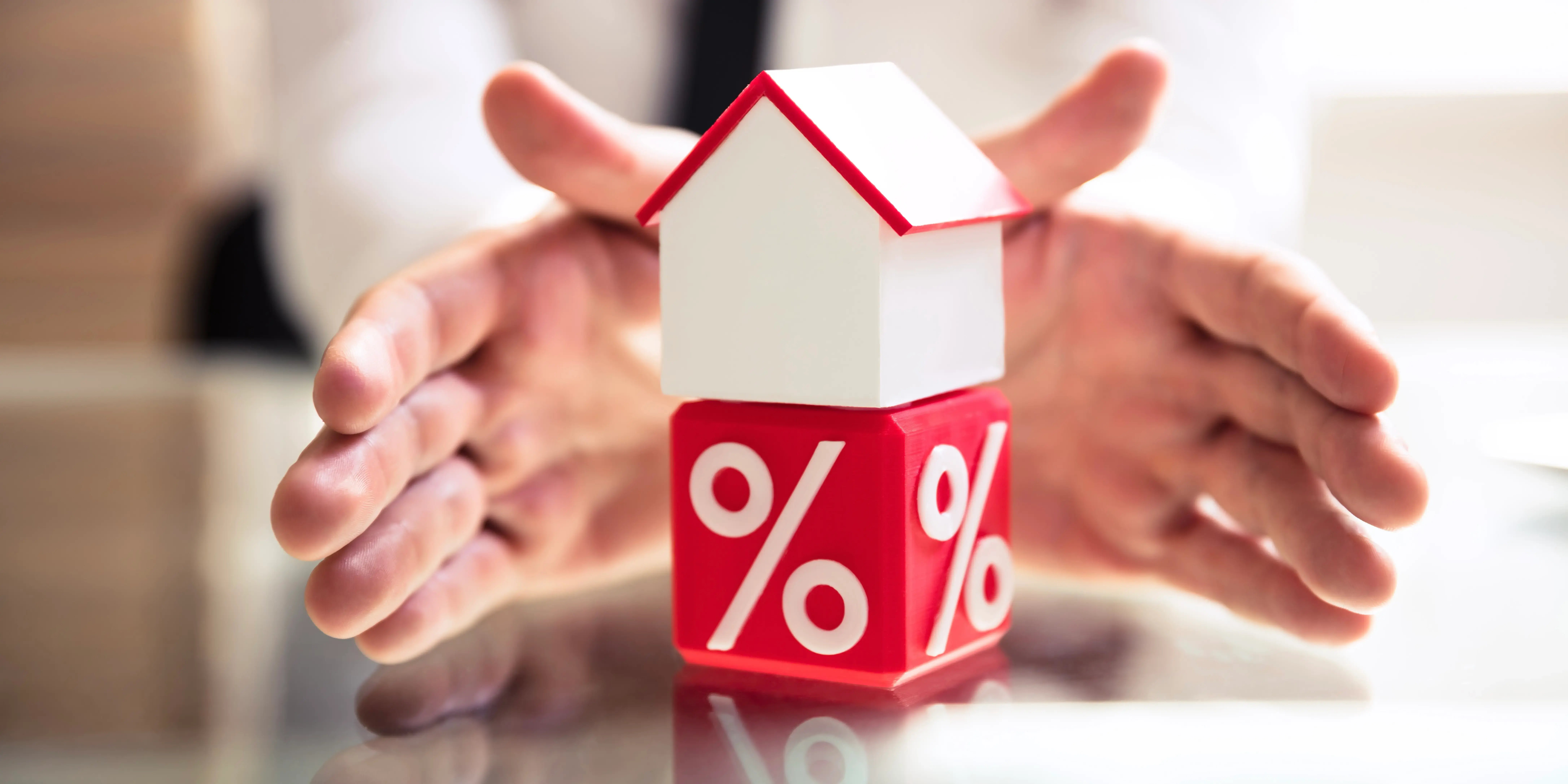 Manos de una persona protegiendo un cubo rojo con el signo de porcentaje y una casa de madera arriba por la temporada de descuentos inmobiliarios