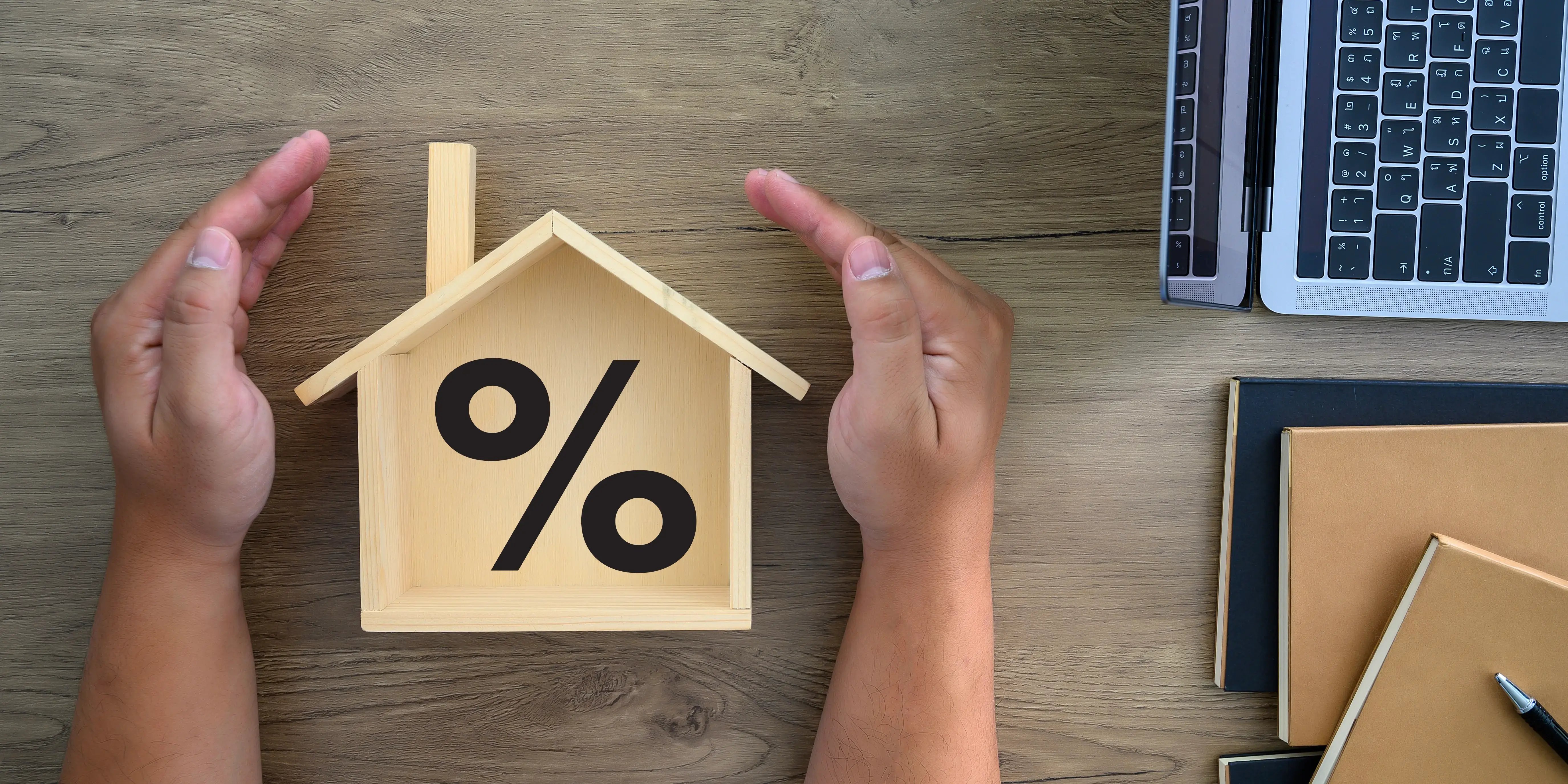 Manos rodeando una casa de madera con un signo de porcentaje adentro como referencia del enganche, uno de los requisitos para crédito hipotecario