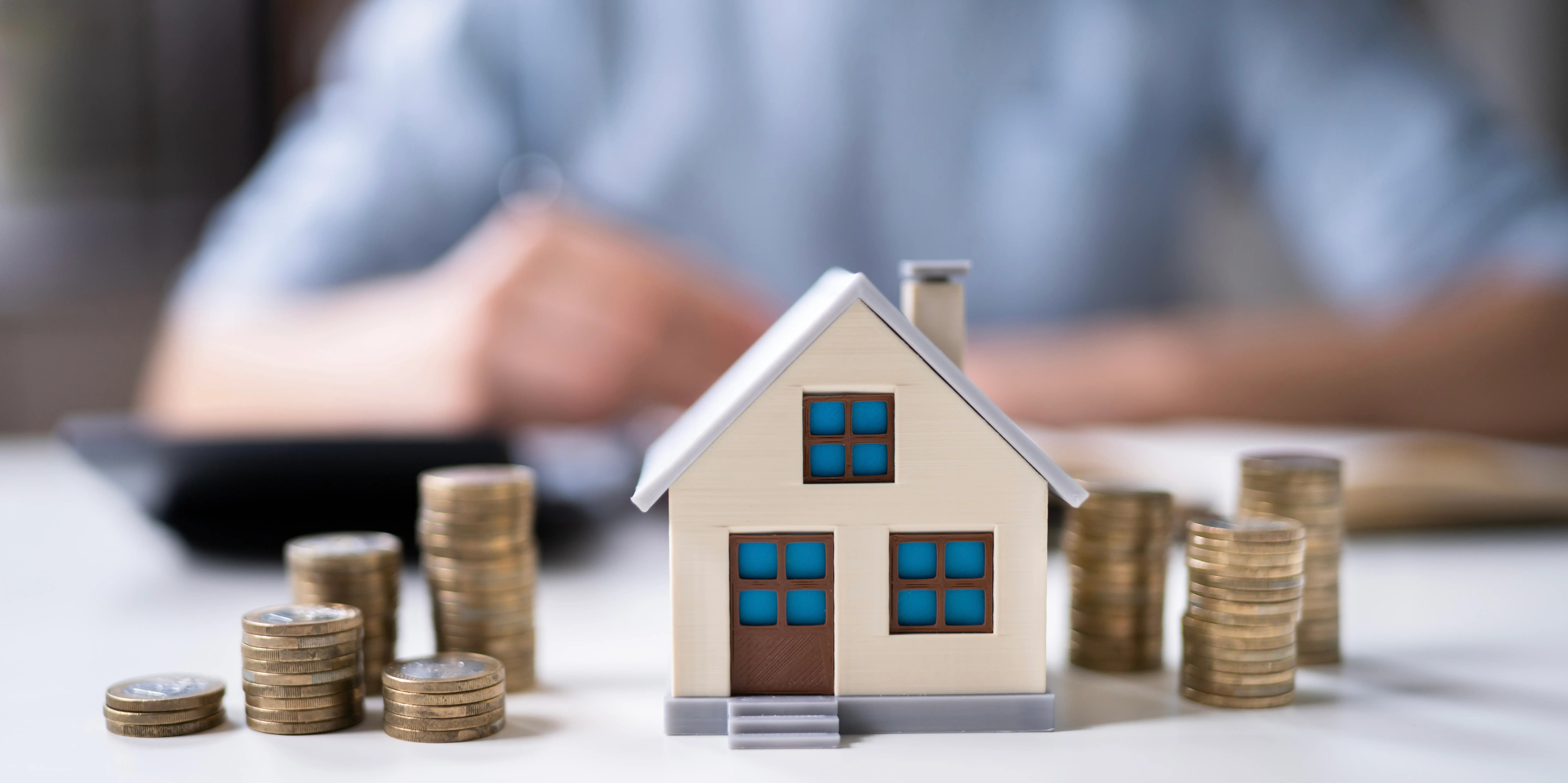 Torres de monedas y en medio una casa de madera, al fondo se ve una persona haciendo cuentas de la renta de la casa que se va acumulando para el encanche y comprar casa