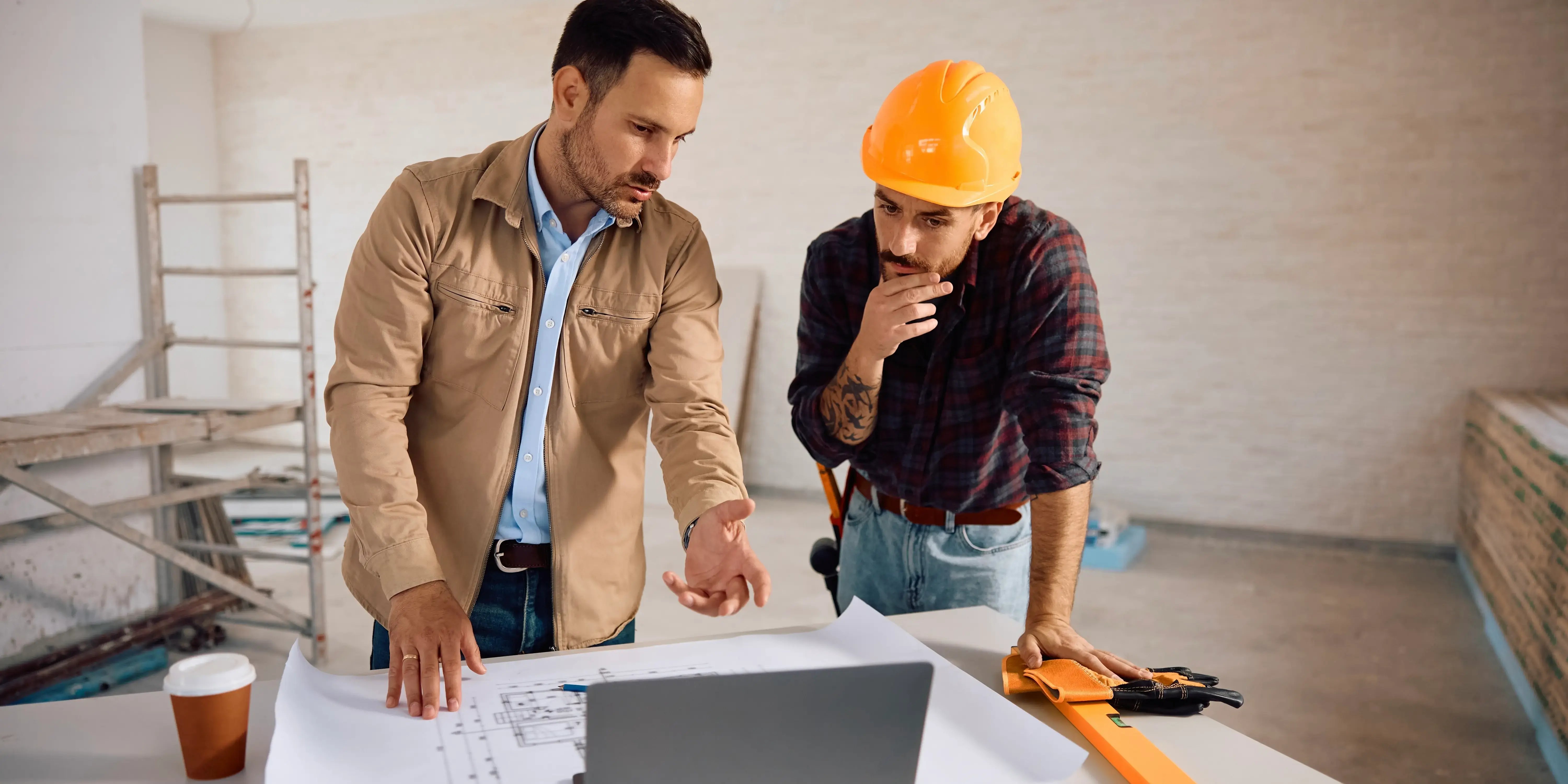 Trabajador de la construcción y su cliente usando la computadora portátil mientras examina los Planes de un desarrollo inmobiliario