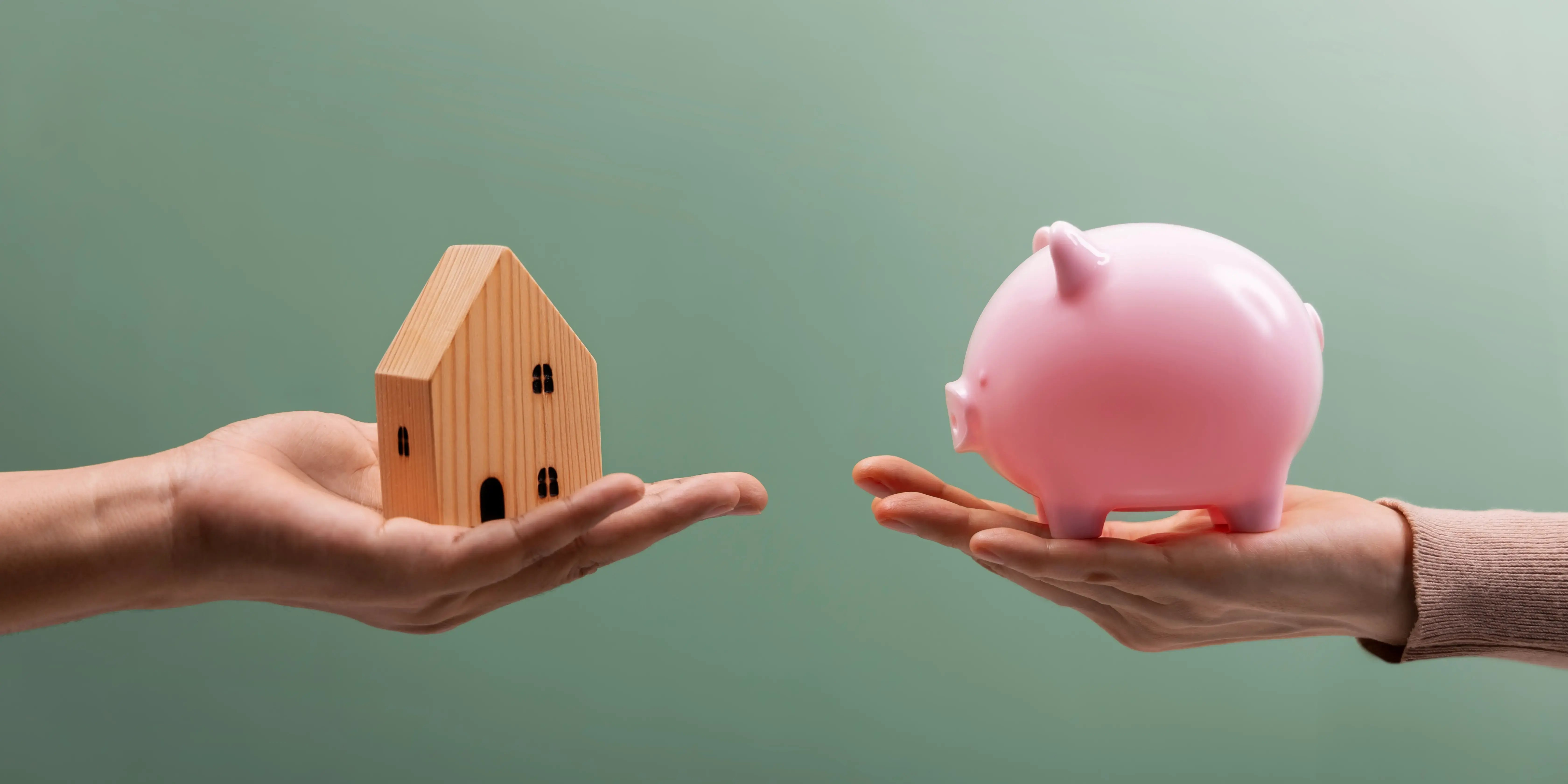 Una mano sosteniendo una casa de medera y enfrente otra mano sosteniendo una alcancía de marrano para comprar una casa sin dinero ahorrado