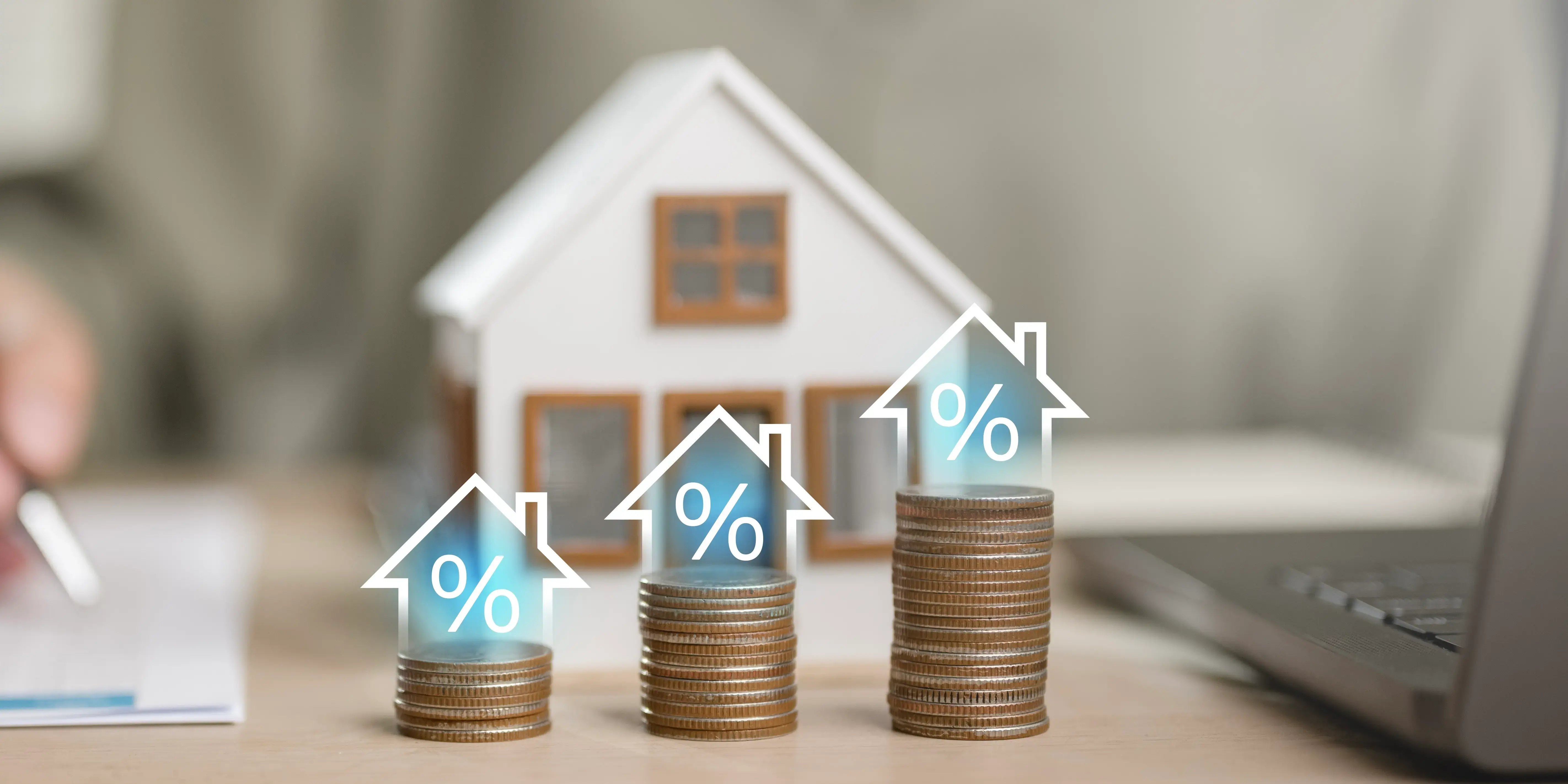 Una persona está sentada en una mesa con tres pila de monedas y en cada una la silueta de una casa y el signo de porcentaje adentro