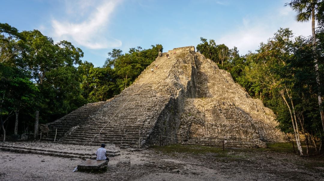 Pirámide de Nohoch Mul, en Cobá. Hay un hombre sentado al frente de la pirámide; al rededor está la selva.