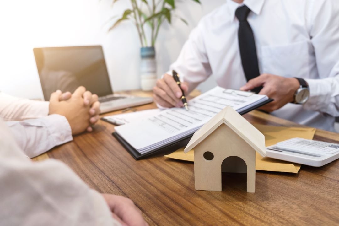 Hombre sujeta documentos legales para una casa, está señalando detalles y en su escritorio hay una casita de madera.