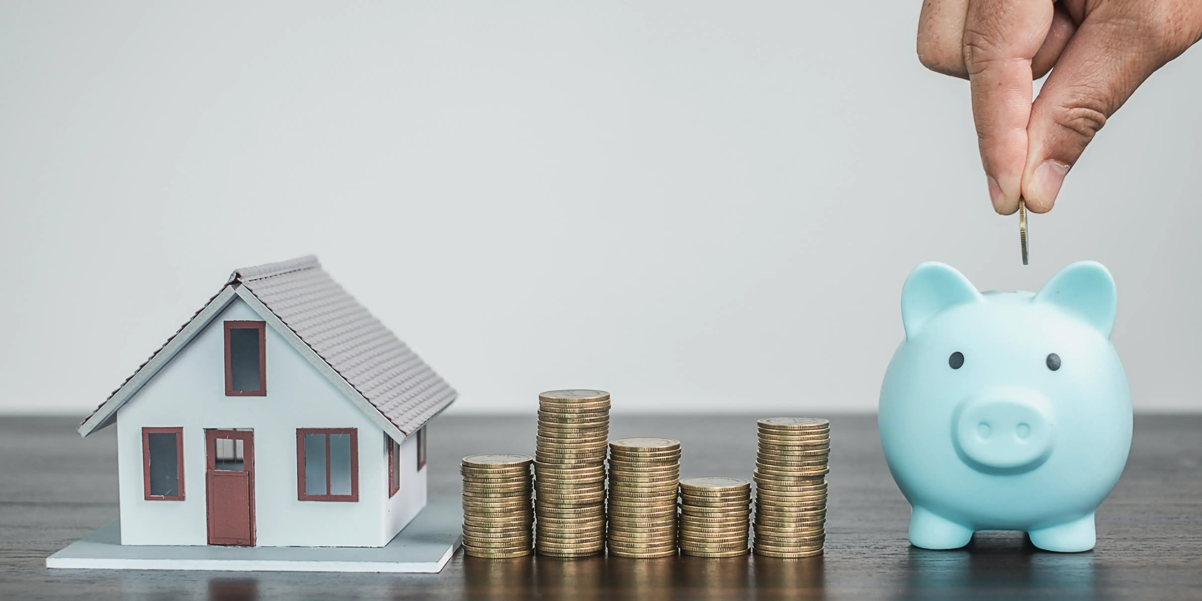 Casa de madera sobre una mesa y una mano guardando monedas en una alcancía como ahorro de la subcuenta de vivienda y AFORE.